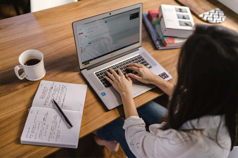 Woman typing on a laptop with handwritten notes and coffee on a wooden desk